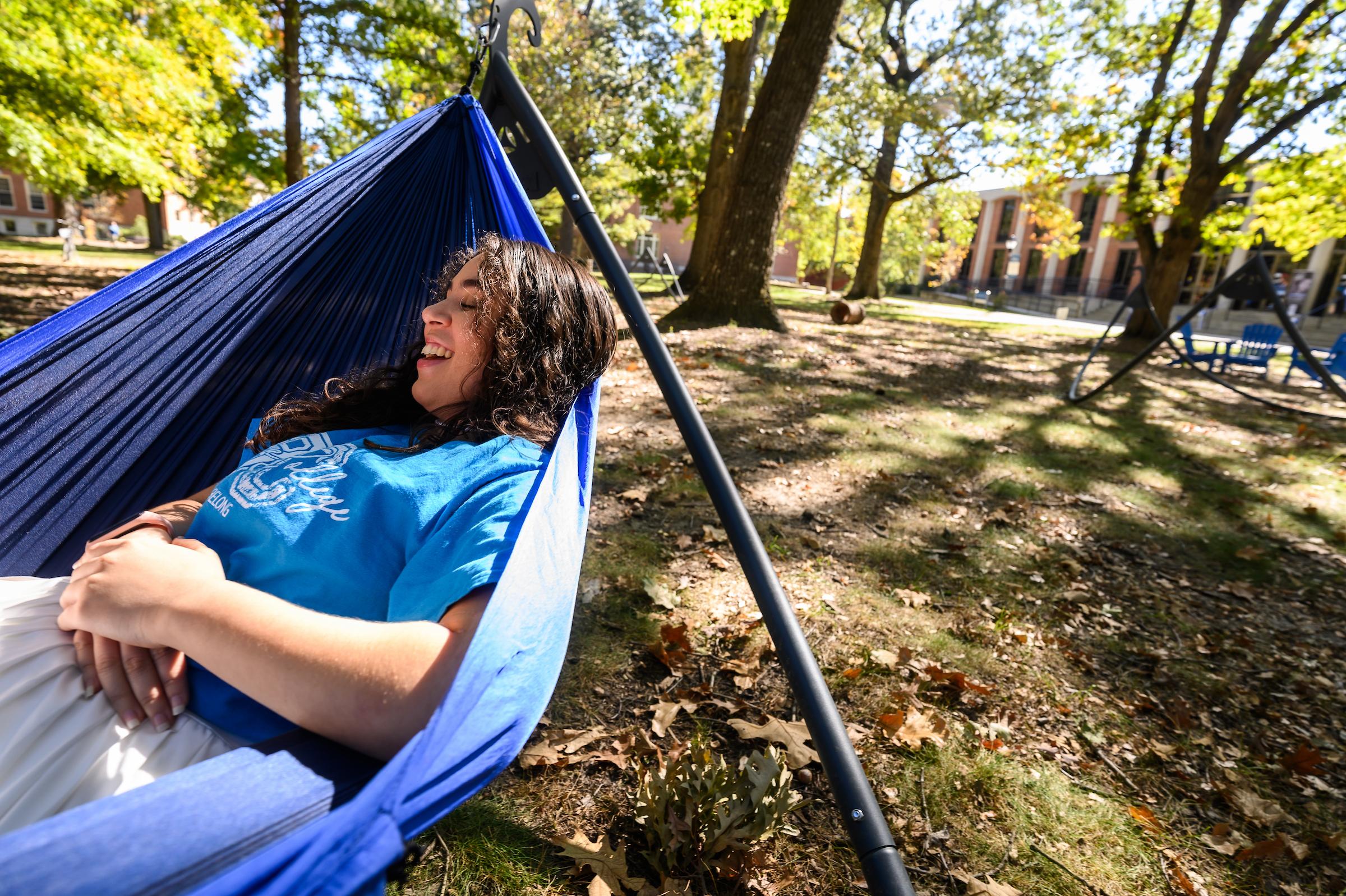 A female student relaxing in a blue Hammock, her eyes and mouth crinkling at the corners as she laughs.