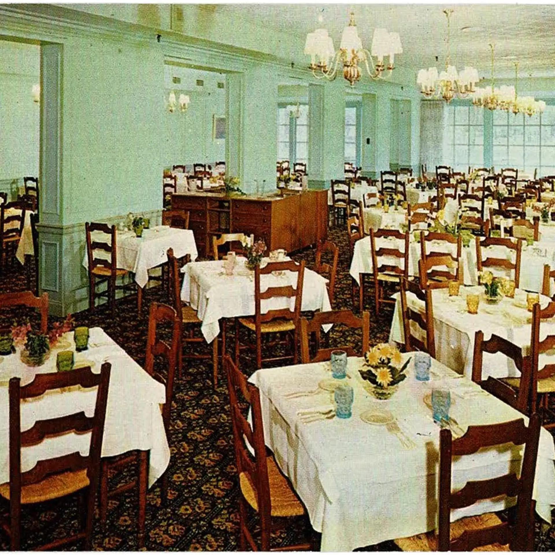 A photo of the Boone Tavern Restaurant from the 1950s, showing lime green walls and an intricately designed carpet flooring. The room is filled with dining chairs and tables covered in tablecloths, cutlery, and dishware.